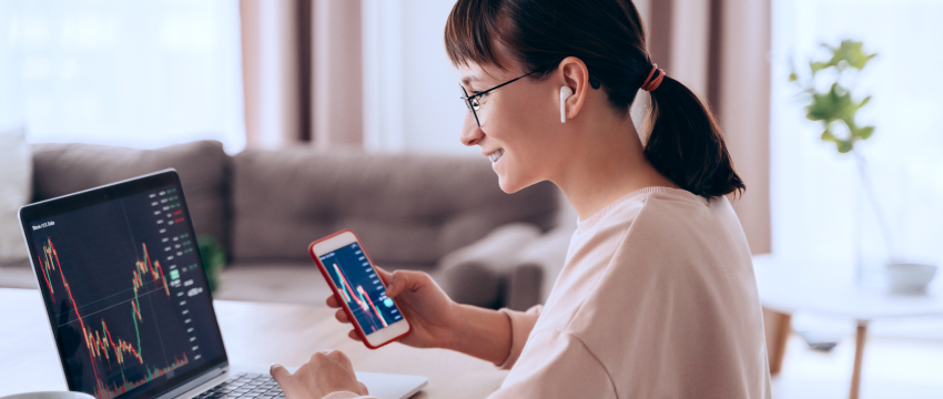 A woman sits at a table with a laptop, using her phone, focused on her work as a forex trader and trading analyst