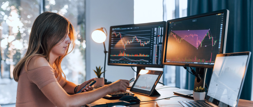 A woman at her desk uses two monitors and a laptop to analyze forex platforms for successful trading.