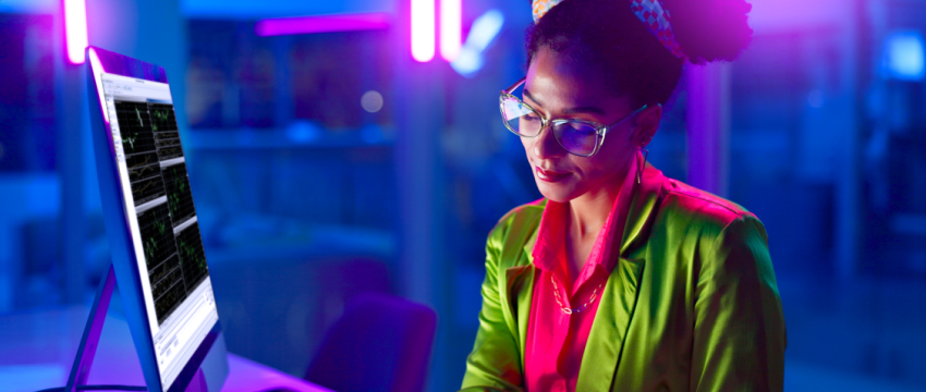A smiling woman wearing glasses and a green shirt is focused on her computer, representing success in trading.